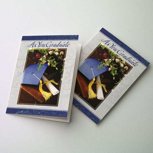 Still life photo of blue mortar board with gold tassel, diploma tied with yellow ribbon surrounded by flowers on an end table; blue marbleized paper border and subtle ornamental design in the gray background.